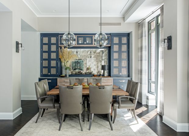 Elegant dining area integrated with a dark blue built-in kitchen or bar wall. A large timber dining table with upholstered chairs sits beneath two glass globe pendant lights, while tall windows with sheer curtains provide abundant natural light. The room features pale walls with decorative ceiling moulding, dark timber flooring, a light area rug, marble-look backsplash, and blue cabinetry with glass-front upper sections.