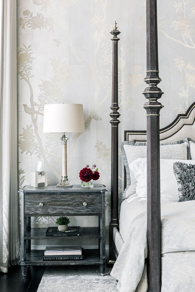 A refined bedroom vignette featuring a tall dark four-poster bed, layered white and grey bedding, a dark wood bedside table with open shelving, and a classic table lamp. Pale botanical patterned wallpaper, full-length curtains, a textured rug, and a small floral arrangement create an elegant, softly monochromatic interior.
