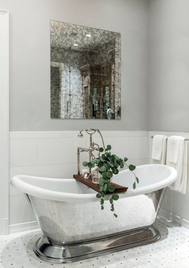 A bright, elegant bathroom featuring a freestanding white slipper bathtub with a polished metal base, exposed vintage-style chrome plumbing, and a wooden bath tray decorated with greenery. White tiled walls, a patterned mirrored panel, soft towels, and a terrazzo-like floor complete the refined traditional interior.