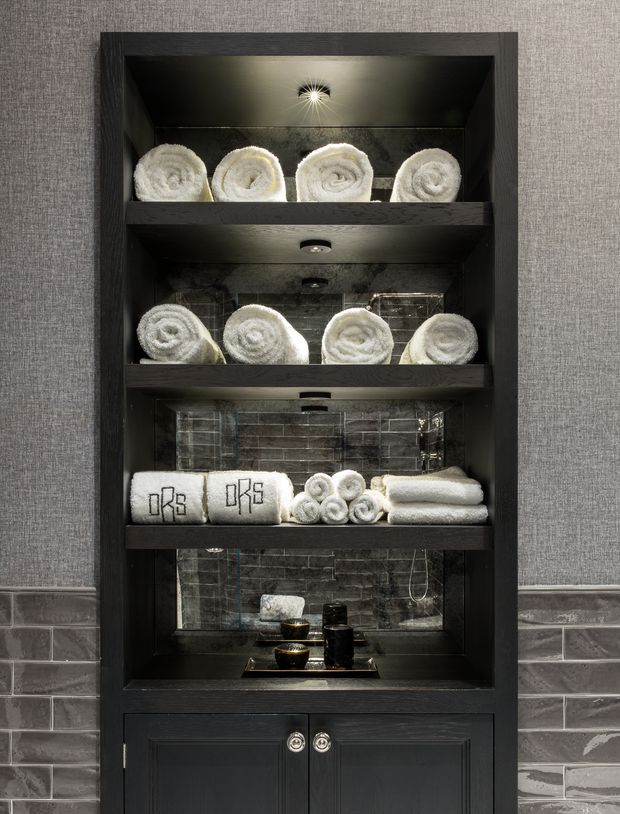 A built-in dark timber storage cabinet in a bathroom, illuminated with integrated spotlights and displaying neatly rolled and folded white towels. The cabinet has open shelving with a reflective dark grey tiled backing and a lower enclosed cupboard with round metal knobs. Surrounding walls combine grey textured wallcovering above and grey brick-pattern tile below, creating a refined, spa-like presentation.