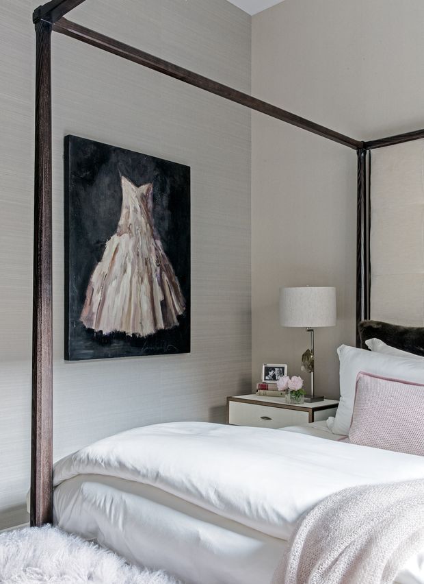 A refined bedroom interior featuring a dark timber four-poster bed, crisp white bedding, a pale upholstered accent pillow, a light timber bedside table, and a table lamp. Neutral textured wallcovering and a monochromatic fashion painting create a calm, elegant atmosphere.