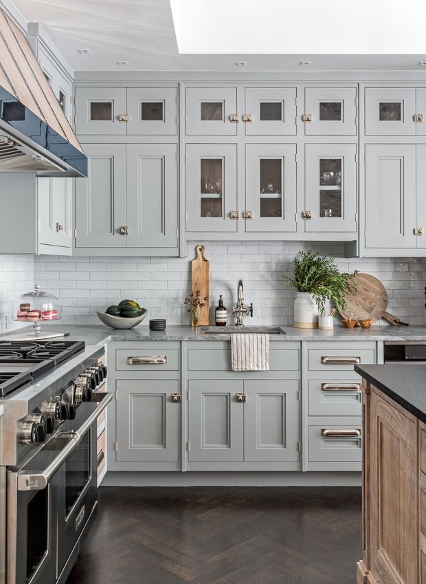 A refined, traditional-style kitchen with grey-blue raised-panel cabinetry, brass hardware, glass-front upper cabinets, a white subway-tile backsplash, dark herringbone timber flooring, and dark stone benchtops. A stainless-steel range and hood occupy the left side, while a farmhouse-style sink, timber cutting boards, greenery, and layered accessories add warmth and texture.