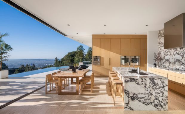 Bright open-plan kitchen and dining area with a large marble-veined island, timber cabinetry, timber dining table and chairs, and floor-to-ceiling openings overlooking a pool terrace and expansive landscape. The space has a flat contemporary ceiling with recessed downlights and a warm, refined material palette.