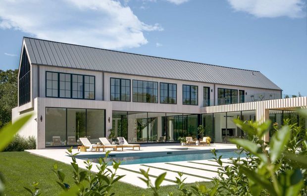 A large contemporary two-storey residence viewed from the pool terrace. The building has a long simple volume, pale vertical cladding, extensive dark-framed glazing, and a pitched standing-seam metal roof. A covered outdoor area extends along the ground floor beside a rectangular swimming pool and paved terrace.