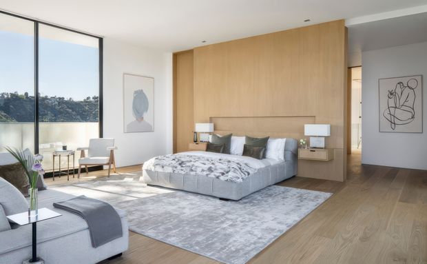 A bright, spacious contemporary bedroom with a low upholstered bed positioned against a full-height light timber feature wall and integrated bedside joinery. Large floor-to-ceiling glazing overlooks a leafy landscape, while pale timber flooring, a large grey patterned rug, neutral bedding, minimal artwork, and streamlined furniture create a calm, refined atmosphere. A partial lounge chair and side table are visible in the foreground.