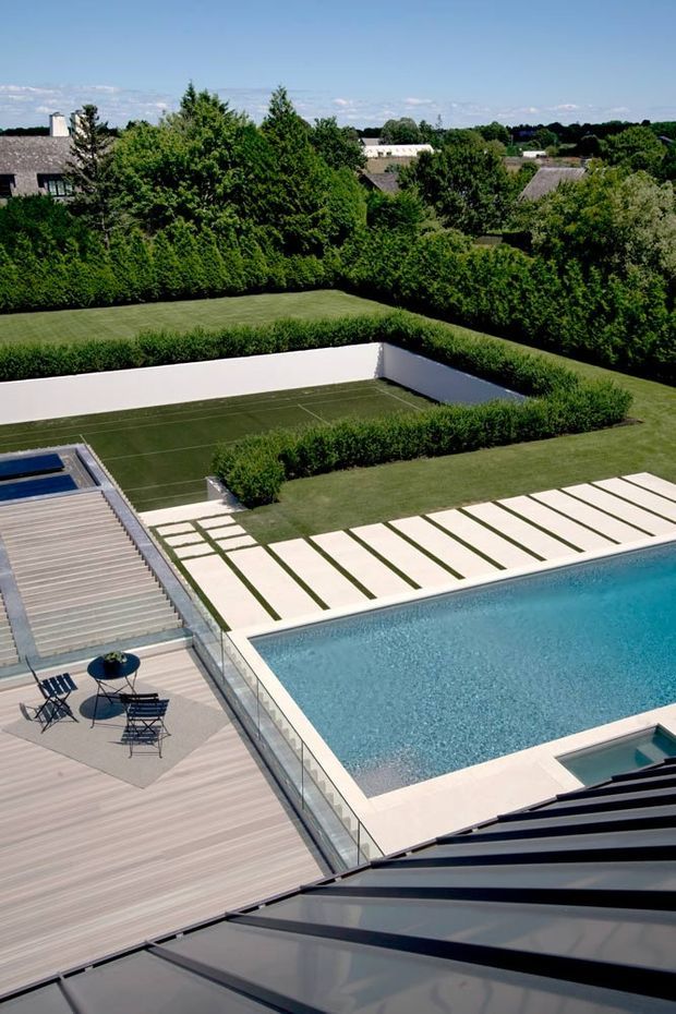 Elevated view over a contemporary landscaped residential property featuring a large rectangular swimming pool, pale paved pool surround with linear grass joints, timber decking, glass balustrades, and a hedge-enclosed tennis court. Dense garden planting and neighbouring pitched-roof homes are visible beyond. The image is bright and summery with clean geometric lines.