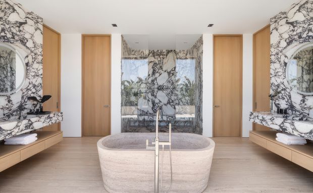 A luxurious, symmetrical bathroom featuring a freestanding oval stone bathtub with a floor-mounted brushed metal filler, twin floating timber vanities, round mirrors, and double timber doors. Dramatic veined marble covers the walls and vanity surfaces, while pale timber flooring and warm wood joinery balance the high-contrast stone. Glazing or a reflective surface behind the bath reveals greenery outside.