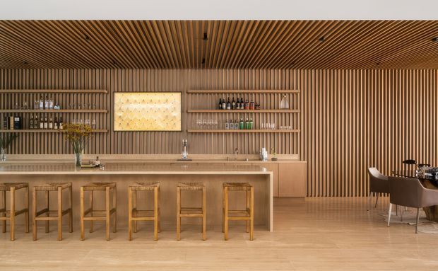 A warm, refined bar and entertaining space dominated by vertical and linear timber slat panelling across the walls and ceiling. A long timber island or bar with multiple wooden stools occupies the foreground, with open shelving displaying glassware and bottles, integrated sinks and a countertop behind it. A small dining area with upholstered chairs is visible to the right. The space has a clean, highly coordinated contemporary timber fit-out with soft integrated lighting.