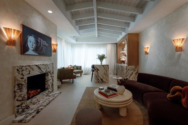 A refined living room with a dramatic marble fireplace and hearth, sculptural rounded coffee table, deep burgundy curved sofa, lounge chairs, and warm wall sconces. The room features textured plaster walls, a pale exposed-beam pitched ceiling, full-height sheer curtains, and a marble-clad built-in bar or display joinery toward the rear. The palette is warm and neutral with dark red accents and art deco-inspired furnishings.
