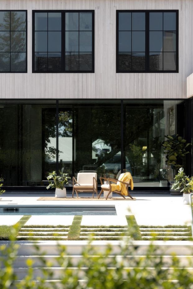 Contemporary two-storey residential facade viewed from a landscaped courtyard. The upper level has pale vertical cladding and large black-framed rectangular windows, while the ground level is largely glazed with dark frames and opens onto a bright paved terrace. Two outdoor lounge chairs and potted plants sit beside a narrow rectangular reflecting pool, with geometric strips of lawn and paving in the foreground.