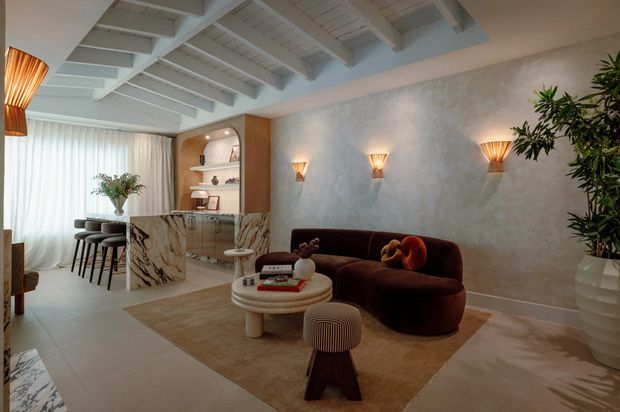 A refined open-plan living room with an integrated home bar or kitchen area. The space features a deep brown curved sofa, round coffee table, textured neutral rug, marble island with dramatic veining, arched timber-toned shelving, sculptural wall sconces, indoor greenery, sheer curtains, and a white-painted exposed-beam ceiling.