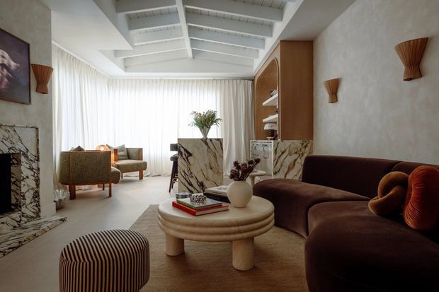 Sophisticated contemporary living room with a large curved dark brown sectional sofa, rounded stone coffee table, striped ottoman, and sculptural decor. The room features dramatic veined marble on the fireplace surround and central partition, warm timber built-in cabinetry, textured plaster walls, sheer full-height curtains, and a white exposed-beam ceiling. A secondary seating area is visible near the glazed wall, creating a bright yet luxurious, softly layered interior.