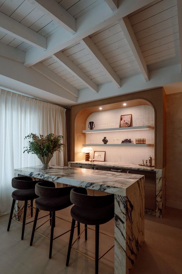 A refined kitchen and bar area with a large veined stone island and four dark upholstered stools. Behind the island is a built-in timber-toned cabinet with a softly arched surround, illuminated ribbed light wall, open display shelves, artwork and integrated lower cabinetry. The room features sheer curtains, warm ambient lighting, and a white painted timber ceiling with exposed beams and rafters.