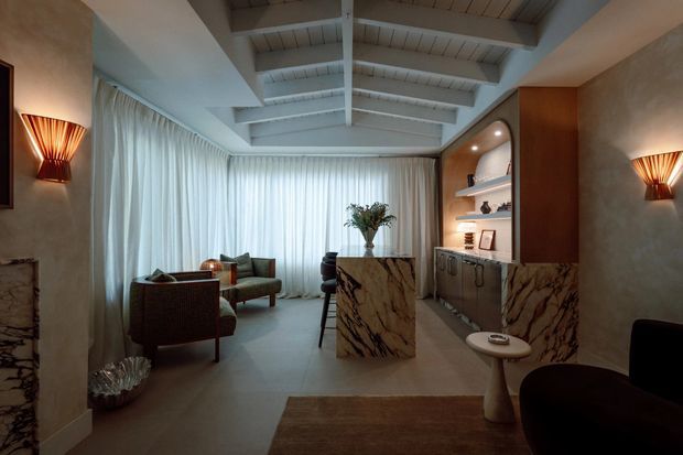 A refined living room with a high exposed timber-beam ceiling, floor-to-ceiling sheer curtains, sculptural lounge seating, and a marble-clad bar or joinery unit. Warm wall sconces, recessed shelf lighting, dark timber cabinetry, and dramatic veined stone create an intimate, luxurious atmosphere.