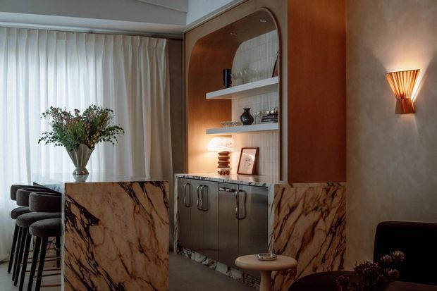 A warm, sophisticated interior corner featuring a built-in bar or display cabinet with rounded timber framing, open shelving, and lower cabinetry clad in dramatic veined marble. Matching marble panels form a partial divider beside a small bar seating area with upholstered stools. Sheer curtains diffuse daylight, while a sculptural wall sconce and carefully styled objects create an intimate, refined atmosphere.
