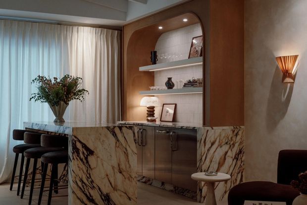 A refined, moody interior featuring a marble-clad bar or kitchen counter in the foreground, dark timber cabinetry, and a softly lit built-in display alcove with curved corners and open shelving. A dining or bar seating area sits beside full-height sheer curtains, while decorative lighting, sculptural stools, and warm wood tones create an elegant, intimate atmosphere.