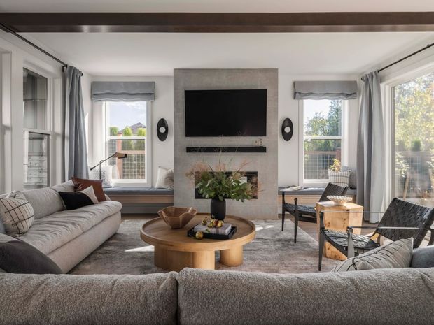 A bright, refined living room with a large sectional sofa, central round timber coffee table, lounge chairs, window seating, and extensive glazing. The room features a contemporary fireplace wall with a wall-mounted television, grey curtains, a textured rug, and a dark exposed ceiling beam.