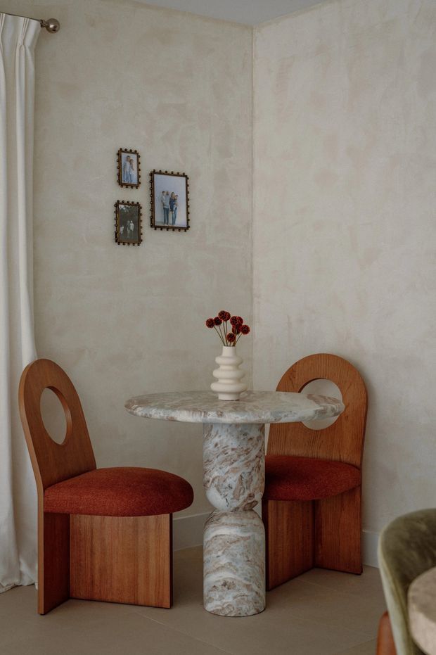 A small, softly lit dining nook with two sculptural wood dining chairs upholstered in rust-red fabric, arranged around a round veined stone pedestal table. The walls have a pale textured plaster finish, with sheer curtains, small framed artwork, and a ceramic vase of dried flowers. A partial upholstered lounge chair is visible in the foreground.