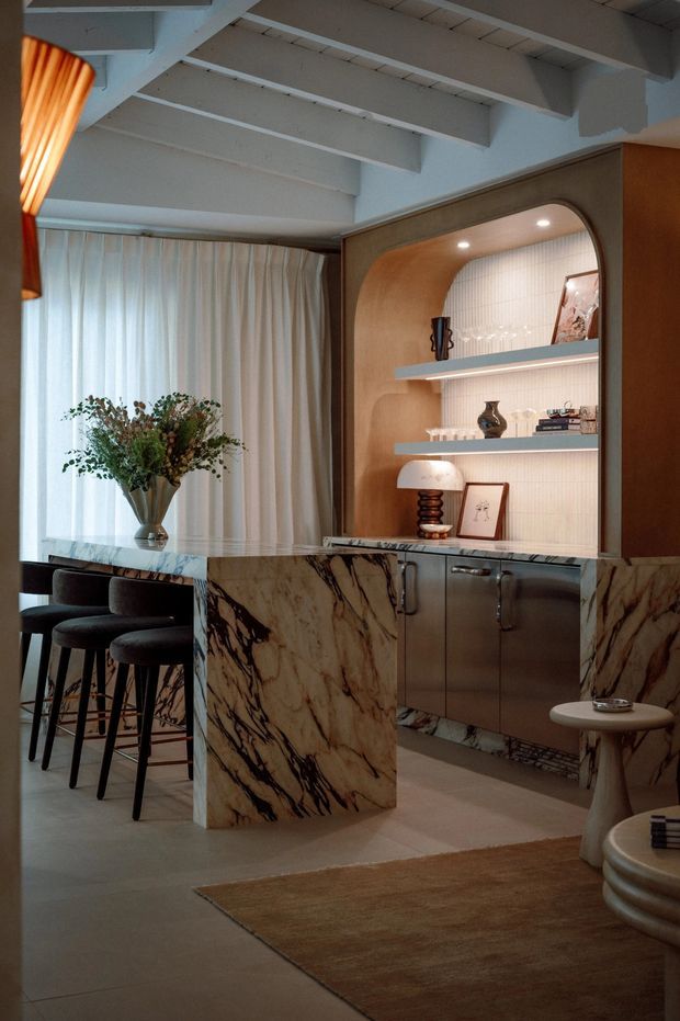 A refined, warm-toned kitchen and bar area with a dramatic veined stone island and matching cabinetry surround. The space features dark bar stools, sheer curtains, built-in arched display shelving with integrated lighting, marble-patterned surfaces, and exposed painted ceiling beams. Decorative objects and a sculptural pendant add a luxurious, curated feel.