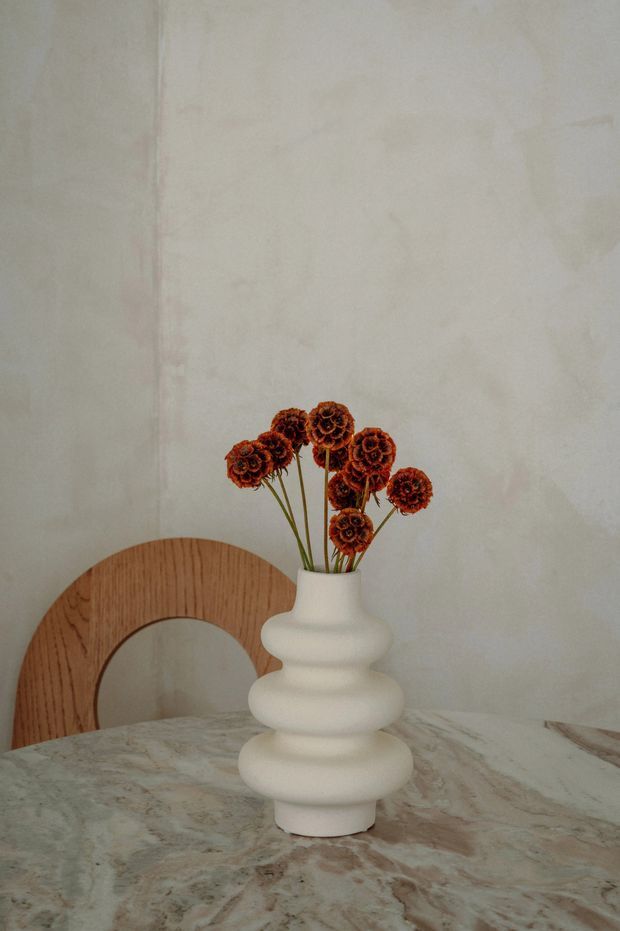 A close-up interior still life featuring a sculptural matte white tiered vase filled with dried rust-coloured flowers, placed on a veined stone tabletop. A curved timber chair is partially visible behind it, set against a softly textured off-white wall. The composition is warm, minimalist, and softly lit.