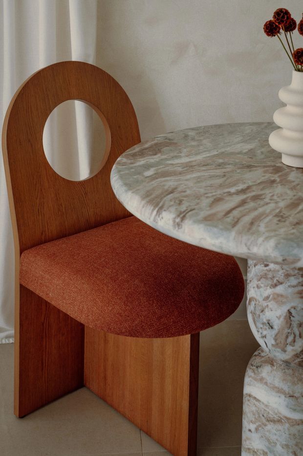 Close-up interior vignette featuring a sculptural wooden dining chair with a circular cutout back and rust-red upholstered seat beside a round, richly veined stone table with a pedestal base. A ribbed white ceramic vase with dried flowers sits on the table. Neutral walls, curtains, and flooring create a warm, refined setting.