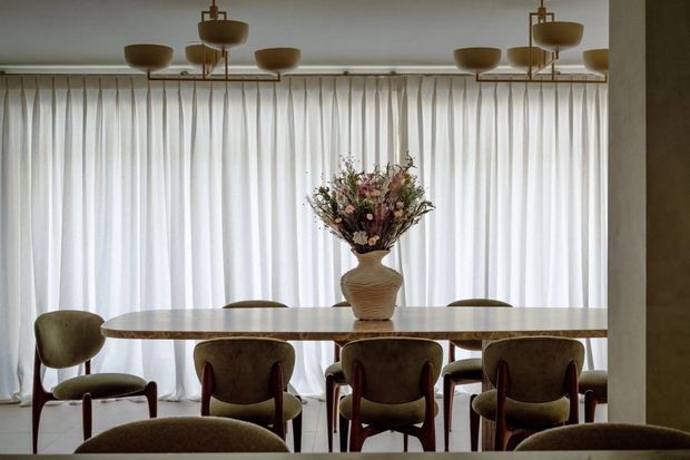 Elegant dining room with a long light-toned dining table surrounded by upholstered mid-century style chairs. Full-height sheer curtains cover the window wall, while a series of brass-toned linear pendant lights hangs above the table. A sculptural vase with dried floral arrangement forms the centerpiece, creating a warm, refined interior.
