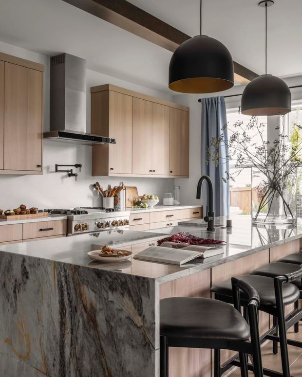 A refined contemporary kitchen with pale timber cabinetry, a large dramatic veined stone island, integrated cooking and prep areas, black pendant lights, and black bar stools. A large glazed opening with blue curtains brings in daylight and overlooks an outdoor area.