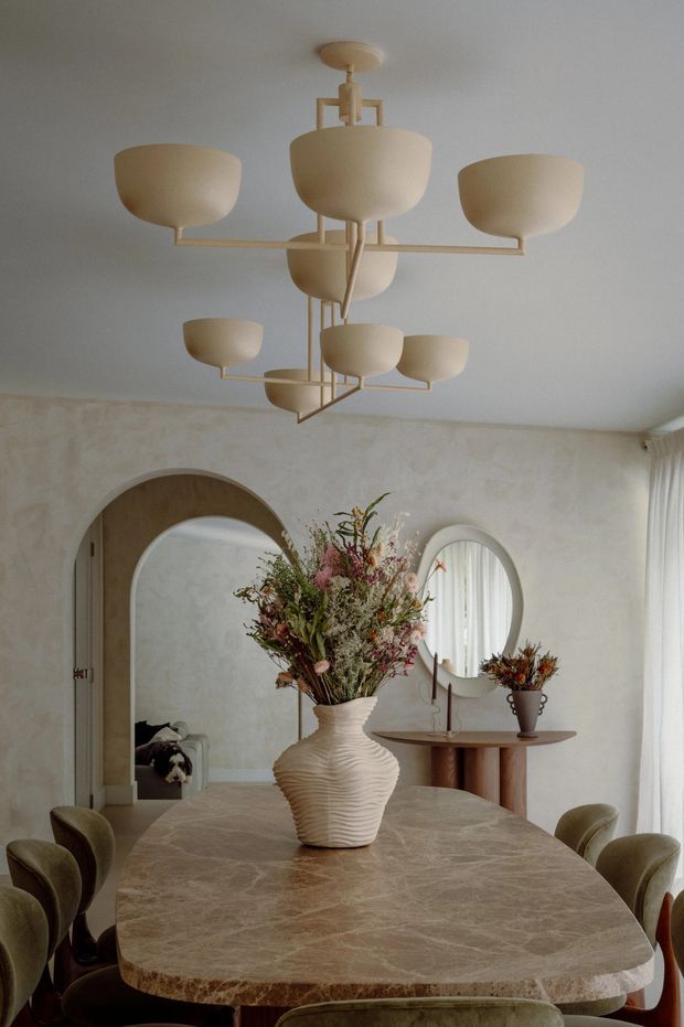 Warm, refined dining room featuring a large rounded rectangular stone dining table, sculptural upholstered dining chairs, and an oversized cream multi-arm pendant light. Textured plaster walls, arched doorways, rounded mirrors, sheer curtains, and organic floral arrangements create a soft contemporary interior with organic, sculptural detailing.
