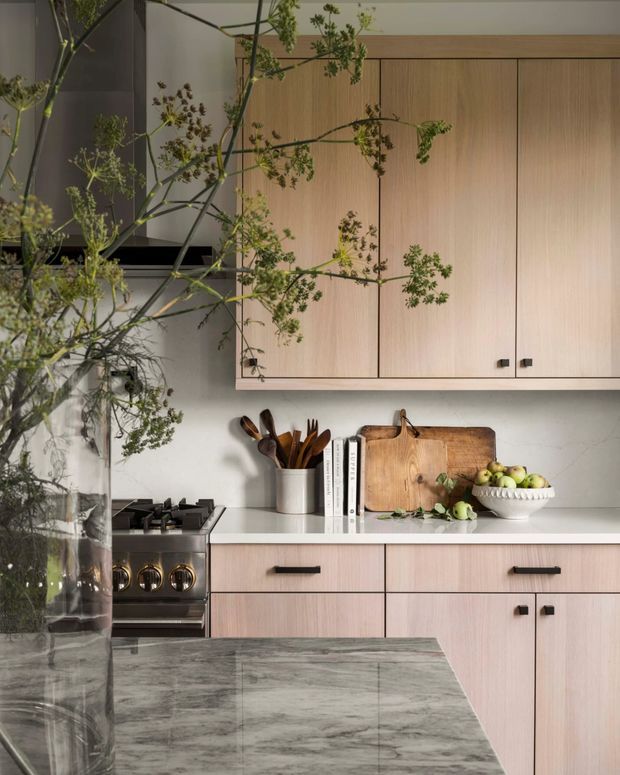 A refined contemporary kitchen with pale wood cabinetry, a light stone benchtop and a darker veined stone island in the foreground. Integrated upper cupboards, dark hardware, a stainless steel freestanding range, timber chopping boards, utensils and a bowl of fruit create a warm, natural composition. Tall leafy branches in a glass vessel partially frame the scene.