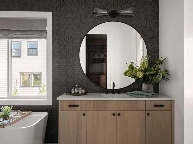 A refined bathroom vanity area with a large round mirror, black textured wall tile, light timber cabinetry, pale stone benchtop, and matte black tapware and hardware. A freestanding bathtub sits beside a window with a Roman blind, while a floral arrangement adds a natural accent. The mirror reflects built-in shelving and an adjacent white interior.