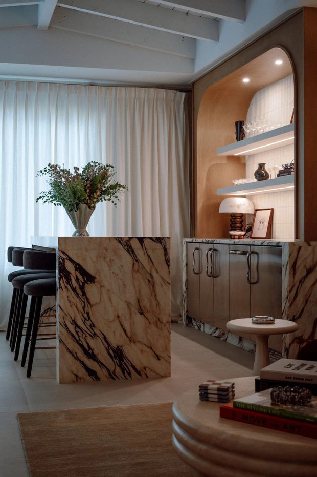 Refined open-plan interior featuring a marble-clad kitchen island and matching low cabinetry, dark upholstered bar stools, a warm timber display niche with integrated lighting and shelving, soft full-height curtains, and a rounded coffee table in the foreground. Exposed painted ceiling beams and layered sculptural furniture create a warm, curated atmosphere.