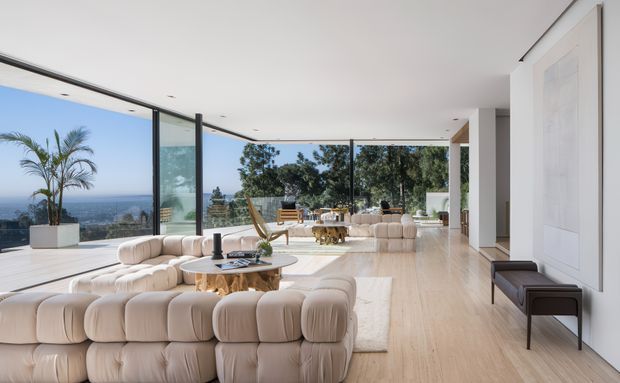 A spacious, light-filled contemporary living room with floor-to-ceiling glazing opening to a panoramic coastal or ocean view. The room features large cream modular sofas, a round wood coffee table, pale timber flooring, white walls, recessed lighting, and a dining area beyond. A broad outdoor terrace and substantial glazing create a strong indoor-outdoor connection.