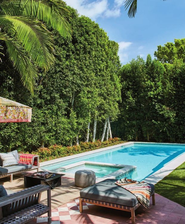 A lush tropical backyard with a rectangular swimming pool and raised square spa, surrounded by dense greenery and palm trees. The pool deck is furnished as an outdoor lounge with cushioned wooden seating, a low coffee table, striped ottomans, and a large patterned outdoor umbrella. The scene is bright and resort-like, with tiled paving and landscaped garden borders.
