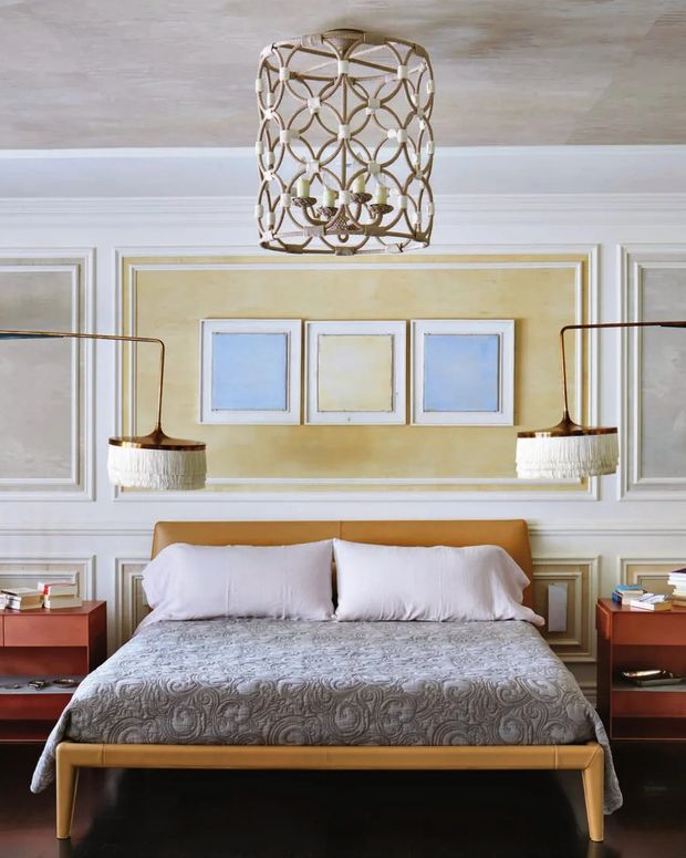A refined bedroom featuring a low-profile upholstered bed with a warm tan headboard and matching footboard, white bedding, and a patterned grey throw. The room has ornate white wall panelling with a muted mustard-yellow inset, symmetrical dark wood bedside tables, sculptural brass-toned wall lamps with fringed shades, and a decorative woven pendant light. Three small framed artworks are arranged above the bed. The overall aesthetic is polished, warm, and traditionally detailed with contemporary furnishings.