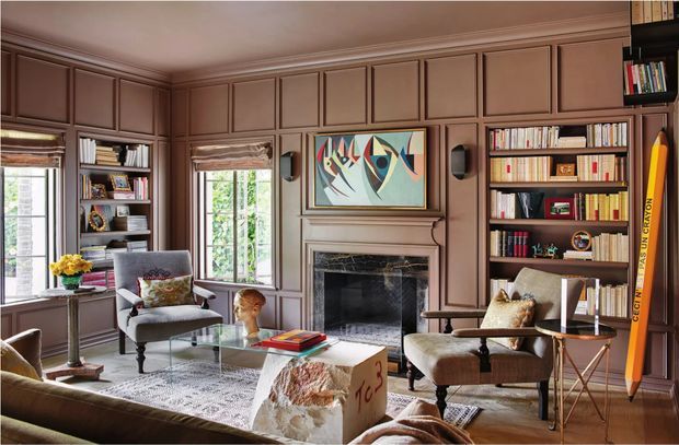 A richly detailed traditional library-style living room with full-height dark wood wall panelling, built-in bookshelves, a central fireplace, tall windows with Roman shades, and layered mid-century furnishings. The room includes lounge seating, a glass coffee table, artwork above the fireplace, and warm natural light.
