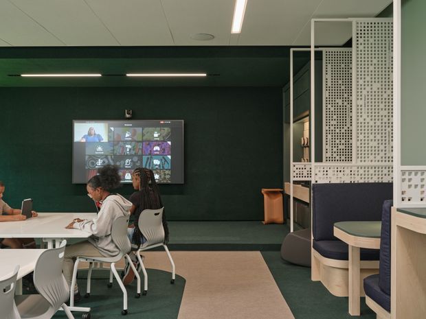 A contemporary collaborative workspace or learning area with people seated at shared tables, a wall-mounted video display, and built-in booth seating. The room features a dark green feature wall, pale timber joinery, perforated white privacy screens, carpeted flooring, and linear ceiling lighting.