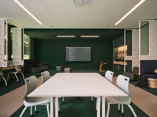 Contemporary collaborative workspace with a large white meeting table and white chairs, dark green walls and carpet, integrated shelving and pinboard-style perforated partitions, linear strip lighting, and a wall-mounted display. Windows on the left bring in views of greenery, while lounge seating and storage occupy the rear and sides.