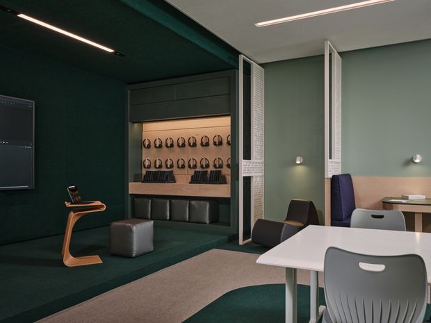 A contemporary media or collaboration room with a dark green built-in lounge and display wall, multiple headphones arranged in illuminated storage, a wall-mounted screen, sculptural side table, and adjacent light-toned meeting workspace separated by perforated folding screens. The room uses integrated linear lighting, muted green walls and ceiling, dark upholstery, and a white table with grey chairs.