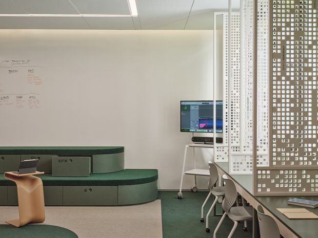 Contemporary open-plan workspace with curved dark green built-in cabinetry and bench seating, a computer workstation, whiteboard, and a long shared desk with chairs. Decorative perforated privacy screens divide the work area, while pale walls, suspended ceiling panels, and muted green and beige finishes create a clean, refined office interior.