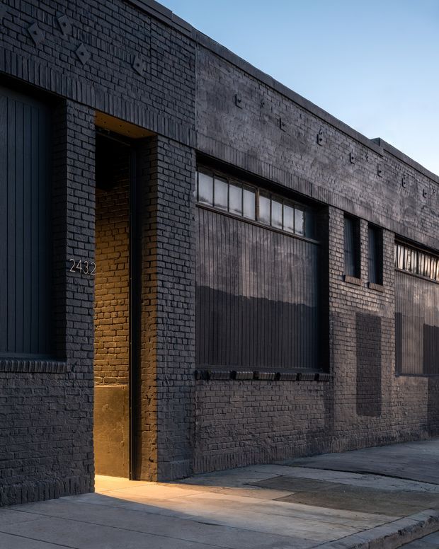 A dark, industrial-style brick building facade viewed from the street. The exterior features painted charcoal brickwork, large corrugated metal-clad window or garage openings, narrow clerestory glazing, a recessed entry with warm lighting, and the number 2432 displayed beside the doorway.