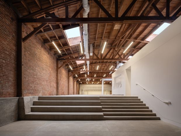 Interior view of a large adaptive-reuse commercial or institutional space, featuring broad concrete steps, exposed red-brick walls, exposed timber roof trusses and rafters, steel structural bracing, linear strip lighting, and visible ductwork. A clean white stair enclosure and handrail contrast with the industrial shell.