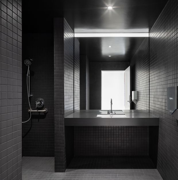 A moody, compact contemporary bathroom with charcoal-black small-format tiled walls, floor and ceiling. A wall-mounted concrete or dark stone vanity with integrated basin and exposed plumbing faces a large mirror, while a concealed shower occupies the left side. A bright doorway or opening at the rear provides strong contrast against the dark tiled interior.