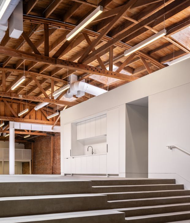 Industrial-style interior with stepped concrete seating or circulation platforms, a compact white kitchenette, exposed timber roof trusses, visible ductwork, brick walls, and linear suspended lighting. The open-plan space combines white contemporary fit-out elements with retained warehouse-like structural details.