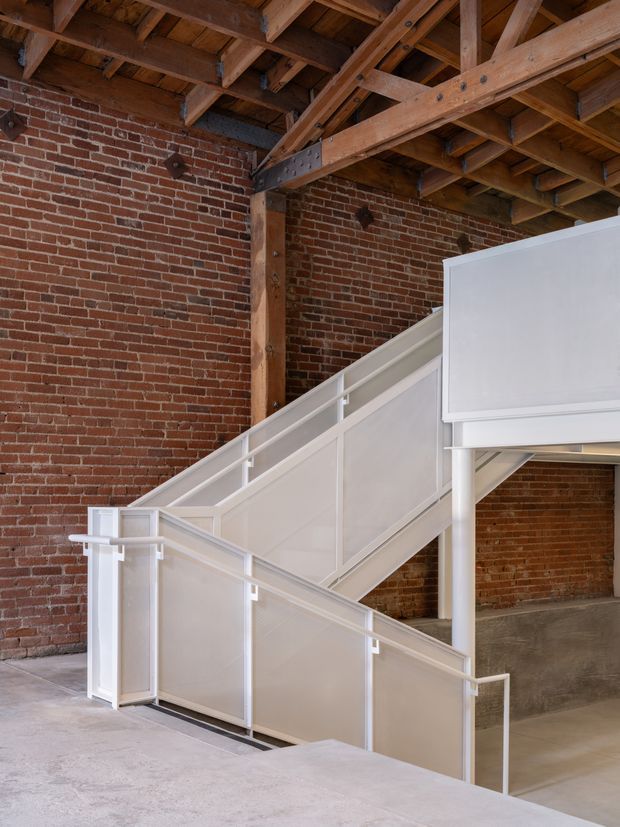 Interior architectural view of a converted industrial space featuring a white-painted metal stair with solid balustrade panels, exposed red brick walls, exposed timber roof trusses and beams, and a raw concrete floor. The composition highlights the stair structure and adaptive industrial character.