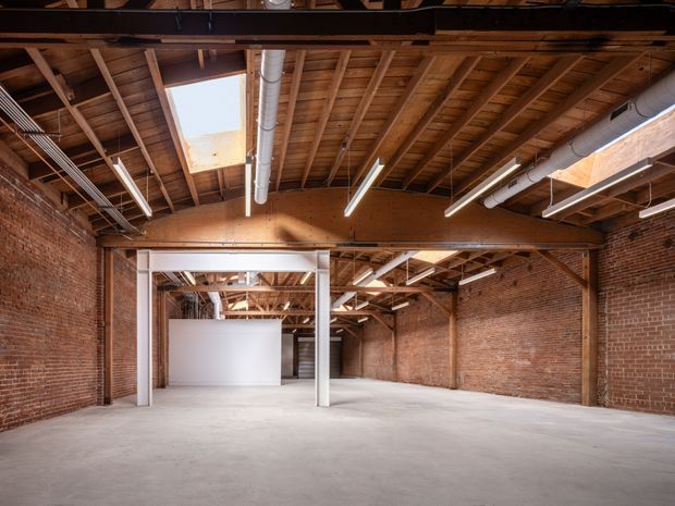 A spacious open-plan industrial interior with exposed red-brick walls, a raw concrete floor, timber roof framing, steel structural beams and columns, visible ductwork, suspended linear lighting, and multiple skylights. The space appears largely empty and adaptable for commercial or creative use.