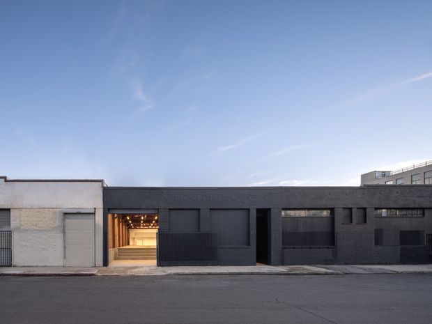 Street-facing view of a low-rise contemporary industrial-style building with a dark charcoal masonry facade, large ribbed metal garage or entry doors, narrow windows, and a warmly illuminated recessed entrance. An adjacent pale rendered section contrasts with the darker main frontage, while a taller urban building is visible in the background.