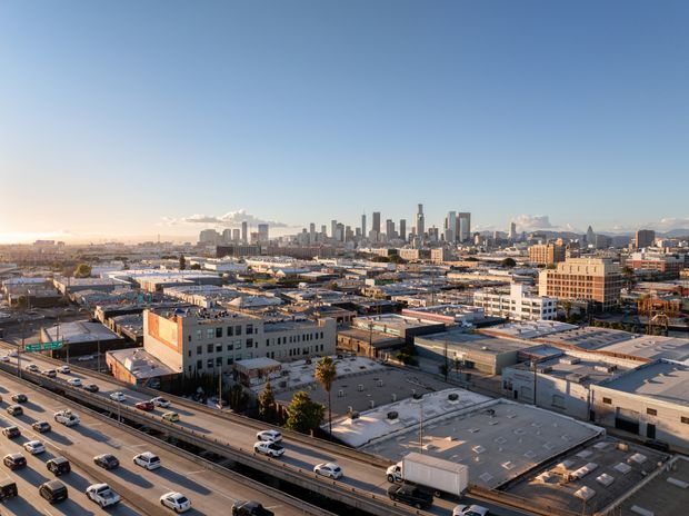 Wide elevated view across a dense urban industrial and commercial district toward a prominent modern city skyline. Flat-roofed warehouses, low-rise commercial buildings, multilane highway traffic, and distant high-rise towers dominate the scene under clear blue sky.