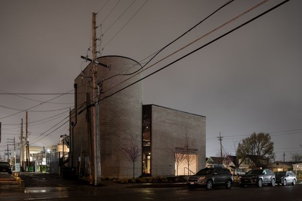 Exterior architectural photograph of a compact contemporary urban building at dusk. The building has a tall curved masonry volume adjoining a simple rectangular wing, with narrow vertical glazing between the forms. Utility poles, overhead wires, parked vehicles, wet pavement, and low-rise surroundings frame the scene.