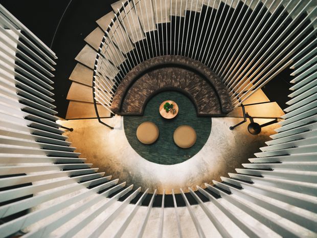 Overhead view of a sculptural circular stairwell or atrium, with pale linear balustrades radiating around a dark circular floor. A curved timber-toned stair or mezzanine edge, two round tan seats, a small central table with greenery, and warm wall lighting create a striking geometric composition.