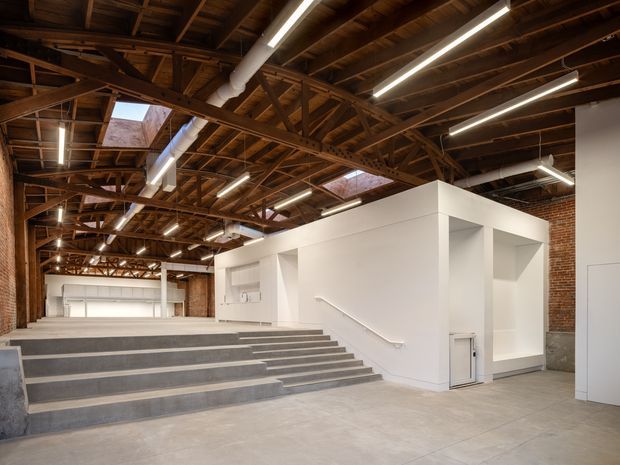 A large adaptive-reuse style interior with exposed timber roof trusses, brick walls, concrete flooring, and extensive linear fluorescent lighting. Broad concrete steps lead to a raised open area, while a crisp white sculptural partition or service volume with recessed openings occupies the right side. Skylights and clerestory-like roof openings bring additional light into the industrial space.